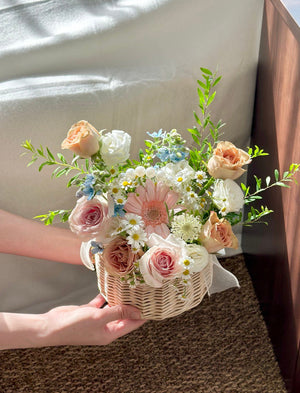 Person holding a small wicker basket filled with a colorful flower arrangement consist of gerbera, quicksand rose, pink rose, snow willow leaf and oxypetalum blue on a carpeted floor.