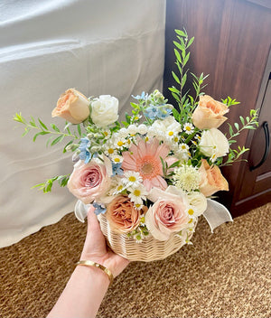 Person holding a small wicker basket filled with a colorful flower arrangement consist of gerbera, quicksand rose, pink rose, snow willow leaf and oxypetalum blue suitable for speedy recovery, house warming and get well soon.