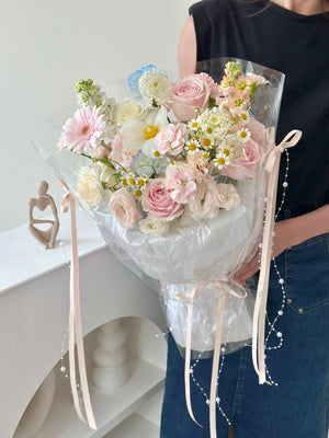 Elegant pastel hand-tied flower bouquet with roses, gerbera, tulip, carnations, chamomile, and trachymene wrapped in clear paper.