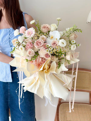 A girl is holding a premium pastel flower bouquet with quicksand roses, white phalaenopsis, and daisy fillers, beautifully wrapped in layered cream and peach paper.