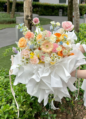 A vibrant pastel flower bouquet with roses, tulip, gerberas, and carnations wrapped in large white ruffled paper, perfect for birthdays or anniversaries.