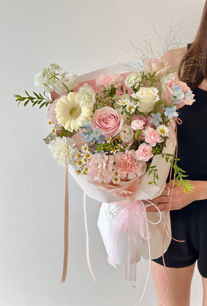 Delicate mix of roses, carnations, and daisies in light pink and white tones, wrapped in white paper with flowing satin ribbons.
