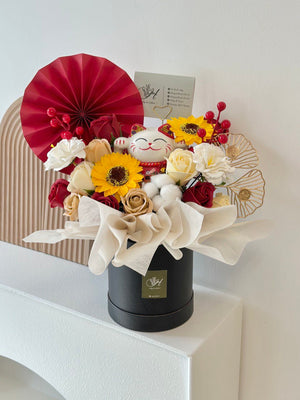A black box with flowers and a red fan, topped with a lucky cat, celebrating a friend's new business opening.