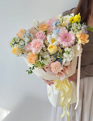 Bouquet of flowers with soft pink, cream, and yellow flowers arranged in a graceful, hand-tied style held by a person against a plain background