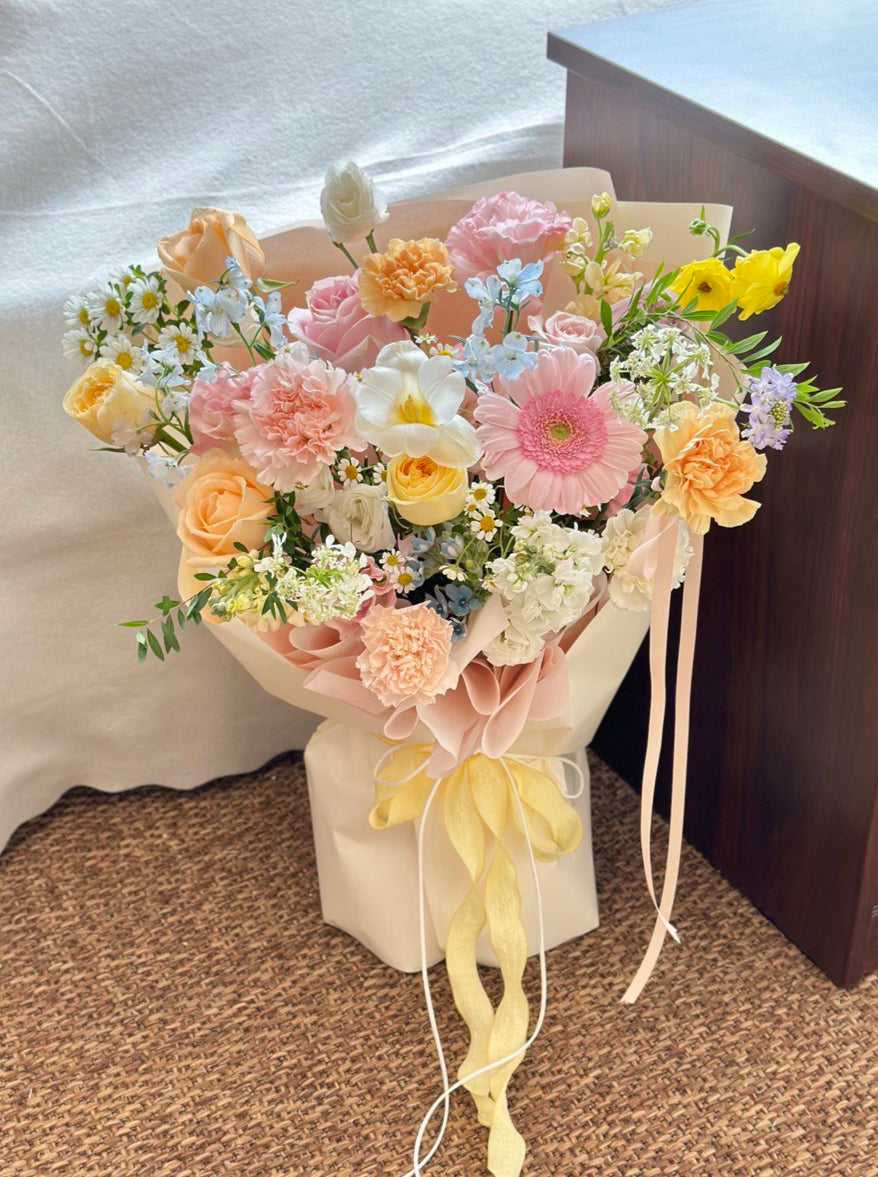 Bouquet of flowers featuring a pastel-toned with pink gerbera, roses, carnations, chamomile, and yellow blooms, wrapped in blush paper with yellow ribbons.