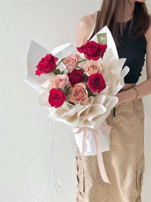 Bouquet of red and cappuccino roses wrapped in white paper held by a person against a plain background.