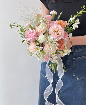 A soft pastel bridal bouquet featuring pink roses, peach carnations, and delicate white daisies tied with a sheer ribbon.