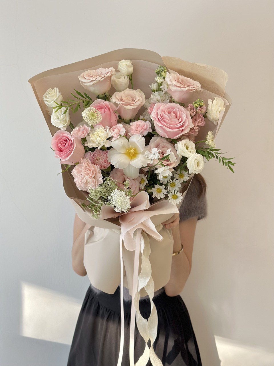 Pastel pink and white floral arrangement featuring roses, carnations, and wildflowers, wrapped in neutral tones with a ribbon.