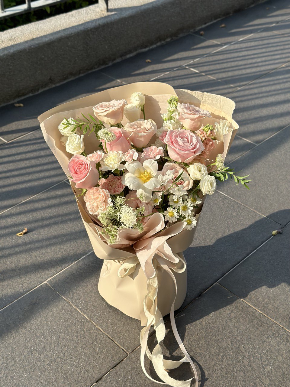 Soft-toned flower bouquet with layered pink roses and white fillers, wrapped in beige and finished with flowing ribbon.