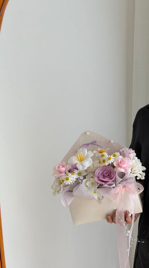 A hand holding a small floral bouquet wrapped in cream paper and sheer pink ribbon, featuring pink roses, white daisies, and a lavender-purple rose, against a plain off-white wall.
