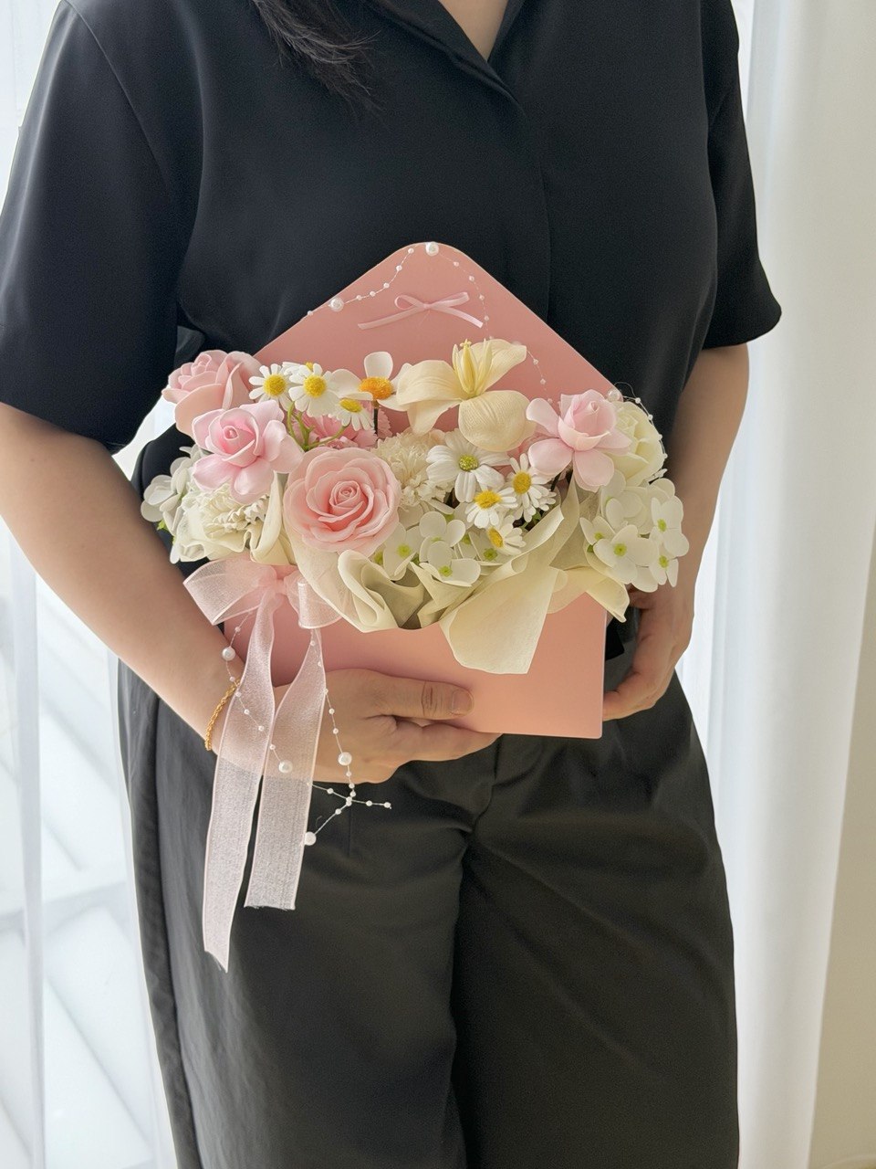 A person in a black short-sleeve shirt holds a pink flower box filled with pink roses, cream flowers, and small white daisies, with pale pink ribbons tied around the front.