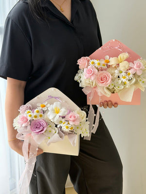 A person wearing a black collared shirt holds two decorative flower arrangements with letter-shaped. One arrangement is cream and the other is light pink, both filled with pink roses, white daisies, and pale yellow flowers with sheer ribbon bows.