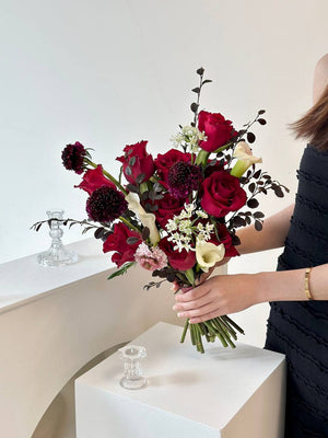 A woman in a black dress holds a wedding bouquet of red roses and calla lilies, preparing for her special day.