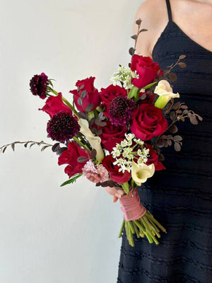 A bride in a black gown holds a vibrant bouquet of red roses mixed calla lilies, preparing for her wedding celebration.