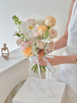 A woman in a white dress holds a bouquet of champagne roses in front of a window on her wedding day.