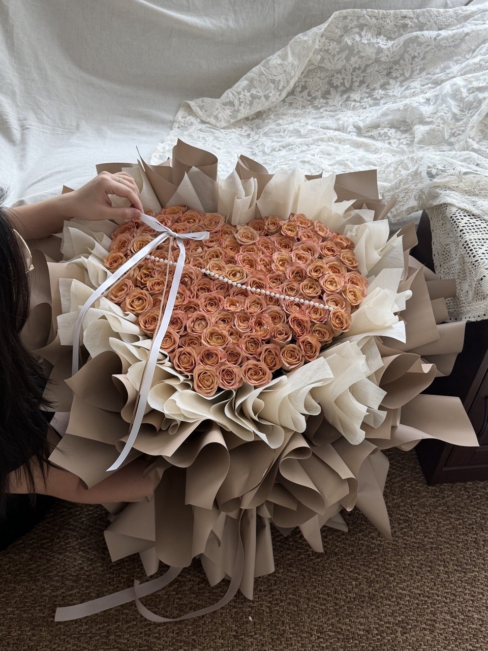 A large heart-shaped bouquet of cappuccino roses wrapped in beige paper and tied with a white ribbon, held by a person whose hands and partial body are visible on the left, with a bed and carpet in the background.