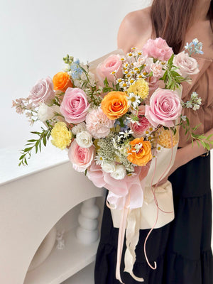 A pastel flower bouquet featuring pink, yellow, and orange roses with soft fillers, wrapped in cream and pink ribbon.