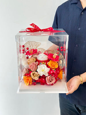 A man presents a clear box with a lucky cat figurine and flowers as a gift for his friend's new business opening.