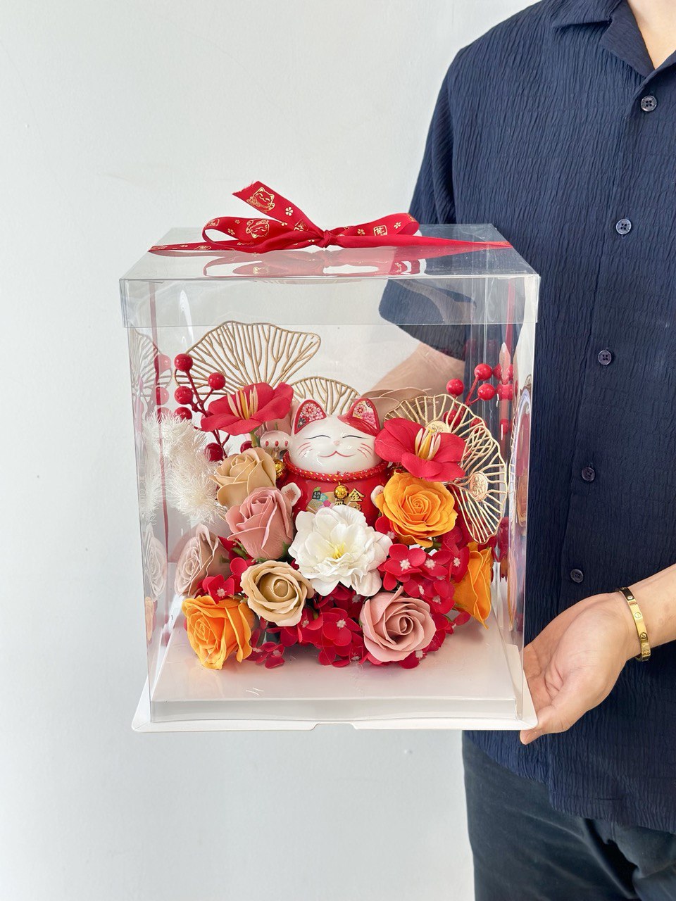 A man presents a clear box with a lucky cat figurine and flowers as a gift for his friend's new business opening.