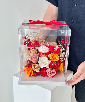 A man is holding a clear box with a lucky cat figurine and flowers, intended as a gift for his friend's business launch at Kuala Lumpur