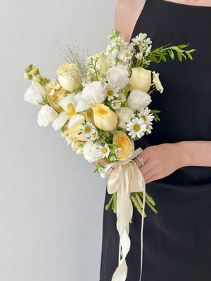 A soft and elegant bridal bouquet featuring buttercup yellow roses, white ranunculus, and hana white flowers, tied with flowing cream ribbons.