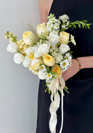 Handheld bridal bouquet with pastel roses, lush white flowers, and whimsical greenery, wrapped in trailing ivory ribbons.