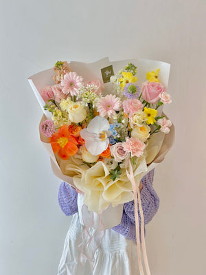 A woman in a purple cardigan presents a vibrant bouquet of flowers at a white table, celebrating her birthday.
