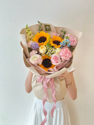 A woman wearing a white dress at her university graduation event while holding a bouquet of sunflowers and pink roses in front of her head