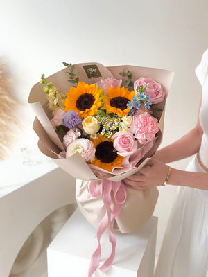 A girl in a white dress proudly holds a bouquet of sunflowers mixed with pink roses, ready for her graduation ceremony.