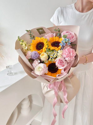 A woman wearing a white dress holds a vibrant bouquet of sunflowers mixed with pink roses, marking her university graduation.
