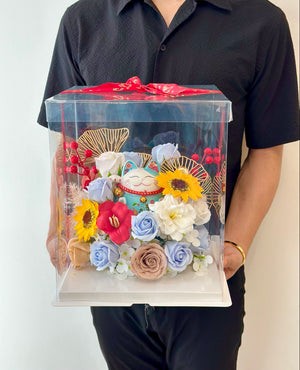 A man with black Shirt is holding a clear box filled with flowers and a blue lucky cat, adorned with a red ribbon, symbolizing a business opening.