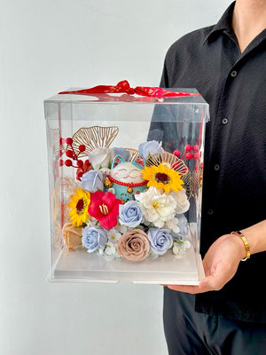 A man is wearing a black shirt is holding a clear box with vibrant flowers and a blue lucky cat, wrapped in a red ribbon, marking a business opening celebration.