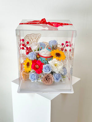 A clear box containing flowers and a blue lucky cat, tied with a red ribbon, celebrating a business opening.