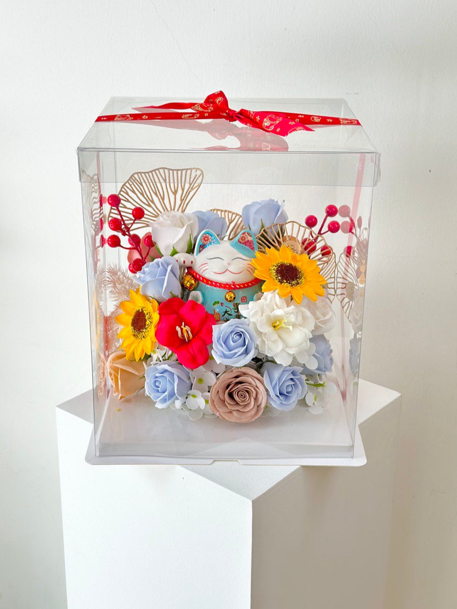 A clear box containing flowers and a blue lucky cat, tied with a red ribbon, celebrating a business opening.