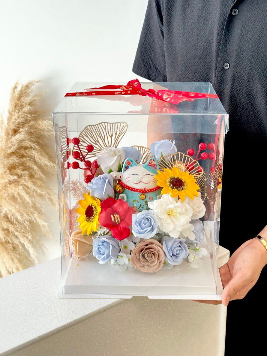 A man with a Gold Band is holding a clear box with vibrant flowers and a blue lucky cat, wrapped in a red ribbon, marking a business opening celebration.