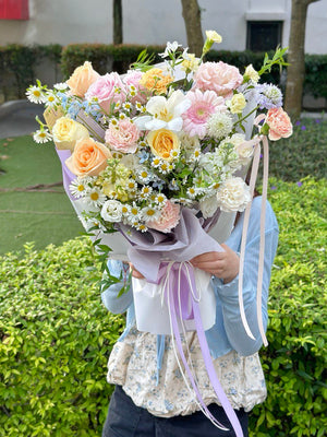Person holding a bouquet of flowers mixed with champagne rose, buttercup rose, white tulip, pink gerbera, chamomile, delphinium blue and other fillersoutdoors with greenery in the background.