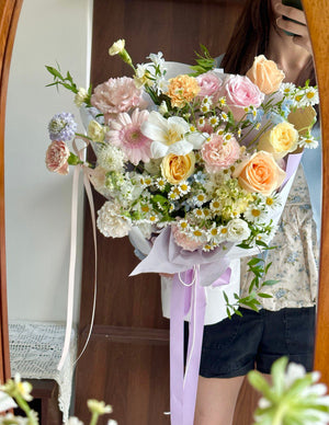 Person holding a bouquet of flowers mixed with champagne rose, buttercup rose, white tulip, pink gerbera, chamomile, delphinium blue and other fillers in front of a mirror.