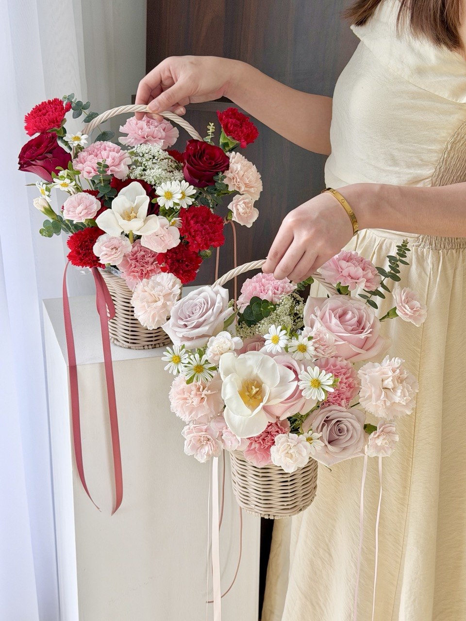 A girl is holding 2 baskets for Mother’s Day flower basket with pink carnations, red roses and white daisies, elegant pastel floral gift Kuala Lumpur same day flower delivery.