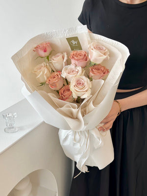 A woman holds a birthday bouquet of roses and cappuccino flowers in her room, with a window in the background.