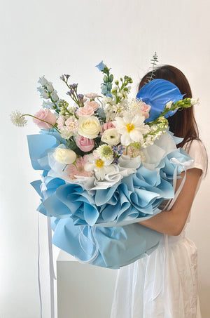 A woman in a white long dress holds a blue and white flower bouquet at her proposal event in Kuala Lumpur.