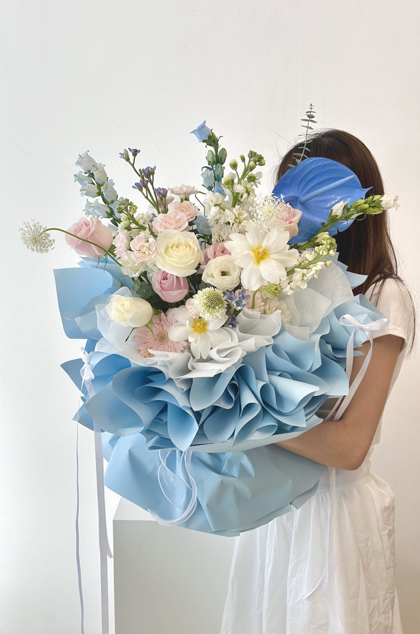 A woman in a white long dress holds a blue and white flower bouquet at her proposal event in Kuala Lumpur.