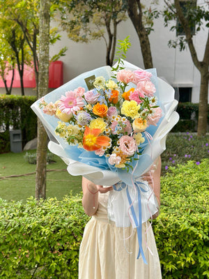 Person holding a bouquet of colorful flowers with pink gerbera, orange tulips, refulgence rose, yellow carnation, buttercup rose and other green fillers outdoors with greenery in the background.