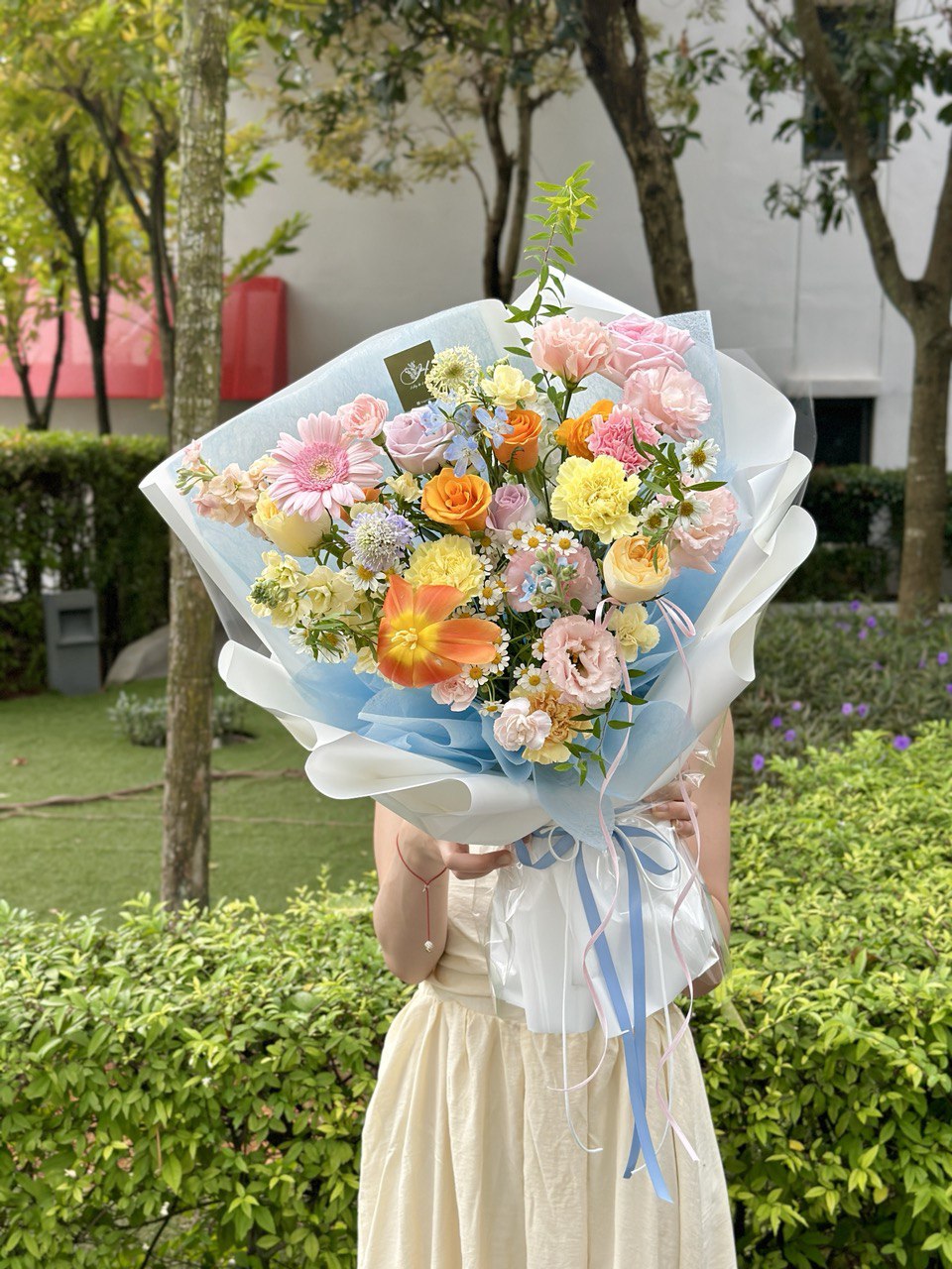 Person holding a bouquet of colorful flowers with pink gerbera, orange tulips, refulgence rose, yellow carnation, buttercup rose and other green fillers outdoors with greenery in the background.