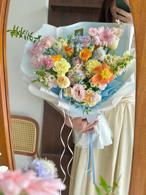 Bouquet of flowers consist of pink gerbera, yellow carnation, orange tulip, refulgence rose, yellow matthiola, lilac purple rose and some fillers held by a person in front of a mirror.