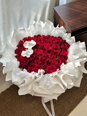 Heart-shaped arrangement of 99 red roses with white orchids on a white surface.