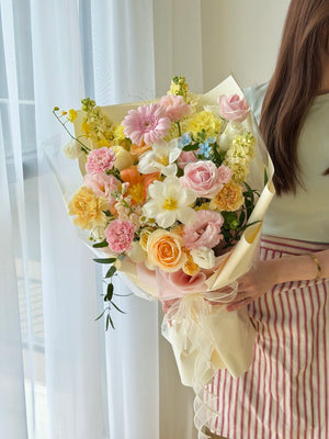 A person holding a large, vibrant bouquet of pastel flowers, including pink roses, pink gerbera, and white tulips, wrapped in cream paper. The person is wearing a light green top and a pink and white striped skirt, standing in front of a window with white sheer curtains.