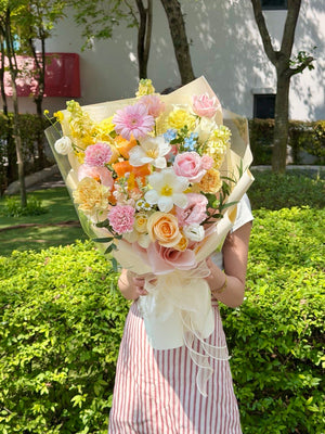 A person outdoors holding a large pastel flower bouquet with pink, yellow, peach, and white blooms wrapped in cream paper. The background shows green hedges and trees.