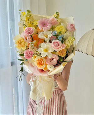 A person holding a large, vibrant bouquet of assorted flowers, including pink roses, dancing lady, and white tulips, wrapped in cream paper. The person is wearing pink and white striped pants, standing in a brightly lit room with a window and a hanging lamp visible in the background.