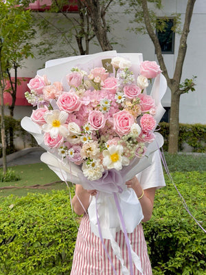 A person outdoors holding a large bouquet of pink roses, pink hydrangea and white flowers wrapped in white paper with a lavender ribbon. The background shows green hedges and trees.
