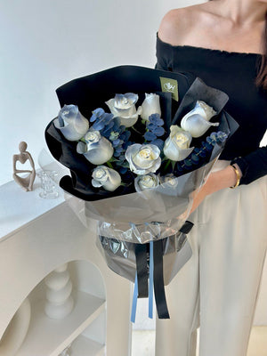 A woman stands in her Kuala Lumpur room, holding a bouquet of white roses, with a cheerful demeanor.